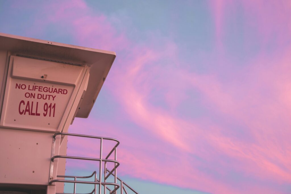 Lifeguard tower with 'No Lifeguard on Duty' sign under a pastel pink sunset sky.